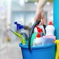 A cleaning woman is standing inside a building holding a blue bucket fulfilled with chemicals and facilities for tidying up in her hand.